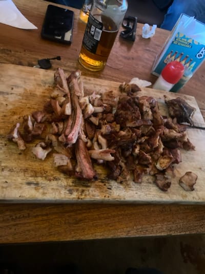 The scene depicts a wooden cutting board laden with various pieces of cooked meat, possibly goat or beef, centrally positioned on a rustic wooden table in Kiaga, Kenya. The meat is cut into irregular shapes; some pieces appear to be bone-in ribs, while others are diced or shredded, exhibiting a color range from reddish-brown to dark brown, consistent with grilling or roasting.
On the table surrounding the cutting board, several items are visible. To the left, a black smartphone rests near a set of black keys. In the background, a partially consumed glass bottle of beer with a label is present. Further to the right, a dispenser for paper napkins or tissues displays text including "REMARKABLE NOT REMAKEABLE" and graphics of bottled beverages. Adjacent to this dispenser are two small condiment bottles, one with a red cap and the other with a green cap. A crumpled white paper is also visible. The wooden table surface shows grain patterns and some wear. Part of a blue garment is visible at the bottom right, suggesting the presence of an individual at the table.