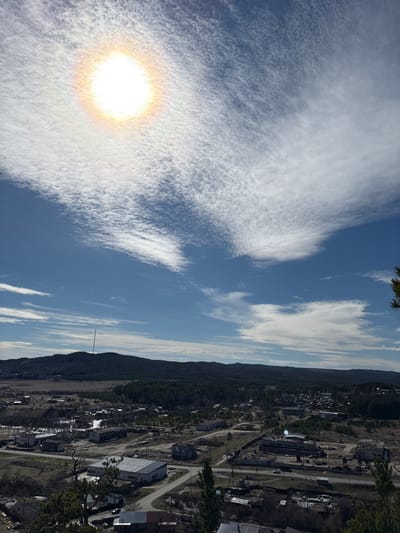 The image shows a view of the city of Невон, Russia, under a bright, sunlit sky with cirrus clouds. The sun is visible through a layer of thin clouds, creating a halo effect. The landscape includes a mix of trees and low-lying hills along the skyline, with a transmission tower visible. The urban area appears to be a small town or industrial settlement with various buildings and roads. The photo is taken from an elevated perspective, possibly a hillside overlooking the town. There are no people immediately visible in the image.