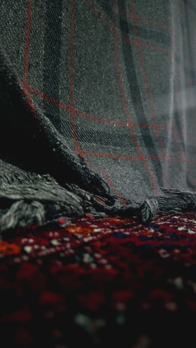 The image depicts a close-up view of a dark grey plaid blanket with thin red lines, draped over a red and blue patterned carpet. The blanket is folded and creased, with frayed edges and tassels visible. The carpet's intricate design is blurred due to the shallow depth of field. The scene is dimly lit, with shadows obscuring details. There are no people or animals present, and no indication of the location in Kaka, Turkmenistan.