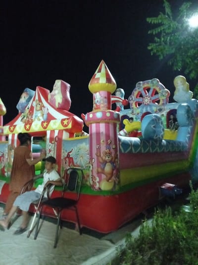 An inflatable play structure, resembling a circus-themed castle with various colorful sections including a Ferris wheel and towers, is set up outdoors at night. The play structure is illuminated by artificial lights, casting a glow on the surrounding area. Two people are visible in the foreground. A woman in a brown dress is standing to the left, facing a young boy sitting on a chair. The boy is wearing a white shirt and light-colored shorts and is looking towards the play structure. A black metal chair with a black seat and back is positioned in front of him. The ground appears to be a light-colored surface, possibly concrete or a mat, with some debris or shadows. Lush green foliage is visible in the lower right corner of the image, suggesting a grassy or garden area. The overall setting is dark, indicating it is nighttime. The atmosphere appears calm, with the people seemingly waiting or observing the play structure. There is no visible text in the image. The location context is Dustlik, Uzbekistan.