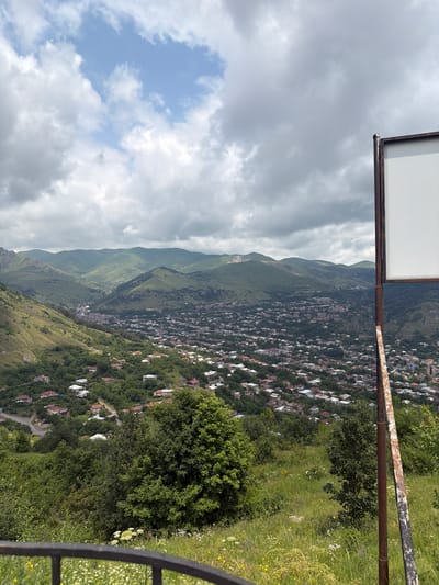 This media file captures a high-angle view of Goris, Armenia, a town nestled within a valley and surrounded by lush green mountains. The scene is dominated by a vast expanse of cloudy sky in the upper left, transitioning to a distant mountain range that appears layered and green. Below the mountains, a densely populated town with numerous houses, many featuring red roofs, sprawls across the hillside. The houses are interspersed with trees, creating a verdant urban landscape. In the foreground, a rusty metal railing and a support pole are visible, suggesting this is a viewpoint or overlook. The weather appears overcast, with the clouds casting a diffused light over the scene. The time of day is indeterminate due to the cloud cover, but the lighting suggests it is daytime. There is no discernible text in the image. The overall impression is one of a scenic and established settlement within a dramatic natural environment.