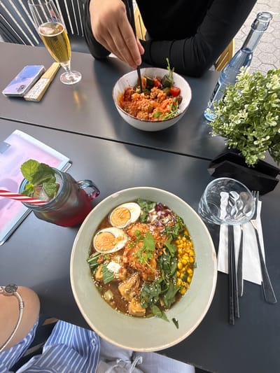 The scene is a top-down view of a table at a restaurant, likely outdoors. The location is indicated to be Aschaffenburg, Germany, though this is not visually evident in the photograph. Two people are present, one on the left wearing black clothing and using chopsticks to eat from a bowl of pasta, and another on the right wearing a blue and white striped shirt and a silver bracelet. On the table, there are two bowls of food: one with pasta and the other with ramen containing corn, eggs, and greens. Additionally, there is a bottle of water, a glass, cutlery and chopsticks, a flower pot, a glass of champagne, a smartphone with a fan on top, and a mason jar filled with a dark liquid with mint leaves and a striped straw. The table itself is dark gray.