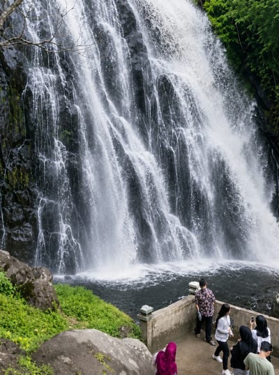 The image shows a wide, multi-tiered waterfall cascading down a dark, rocky cliff face covered in patches of green moss and vegetation. The water is white and frothy in places, creating a dramatic effect. At the base of the waterfall, dark water churns and pools. In the foreground, to the right, a concrete viewing platform runs along the edge of the water. Several people are present on this platform, observing the waterfall.

There are five individuals visible on the platform:
* A person wearing a floral-patterned shirt and dark pants stands facing away from the camera, looking towards the waterfall.
* A woman in a white and black striped shirt and black pants is walking away from the camera.
* Another woman, wearing a white shirt and black pants, is holding a smartphone and appears to be taking a picture.
* A person in a black hijab and white shirt is also on the platform.
* Partially visible at the bottom of the platform is a person wearing a pink hijab, facing away from the camera.
* At the very bottom right of the image, a person wearing a green t-shirt is visible.

The environment includes large rocks and lush green grass on the left side of the image, leading up to the waterfall. Trees with green leaves are visible at the top right of the frame, bordering the waterfall. The overall scene depicts a natural landscape with visitors enjoying the spectacle of the waterfall. The location is identified as Sosor Dolok, Indonesia.