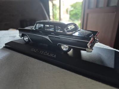A black, 1:43 scale model of a GAZ-13 Chaika limousine is displayed on a black display stand. The model is positioned at an angle, with the rear of the car facing towards the right side of the frame. The car is black and has chrome detailing on the grille, trim, and wheels. The words "GAZ - 13 ČAJKA" are inscribed on the front of the display stand in silver lettering. The background is blurred, suggesting an outdoor setting with greenery visible through what appears to be a window or doorway. The lighting suggests it is daytime. The image was taken in Iskrets, Bulgaria.