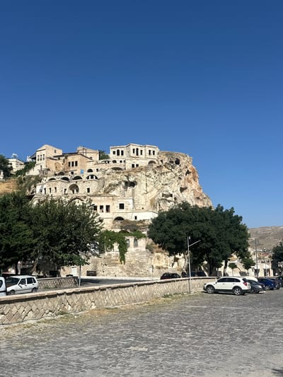 In Ürgüp, Turkey, a view captures a cluster of historic stone buildings carved into and built upon a rocky hillside under a clear blue sky. The structures exhibit a unique architectural style, with multiple levels and arched openings, some appearing to be cave dwellings integrated with more conventional masonry. Green trees with dense foliage are visible in the foreground, framing the hillside dwellings. A cobblestone area in the lower part of the image is partially occupied by several parked cars, including a white SUV, a dark sedan, and a blue car. A stone wall with a diamond pattern runs horizontally across the mid-ground, separating the cobblestone area from the base of the hillside. The overall scene depicts a blend of natural landscape and historical human habitation in a town setting.