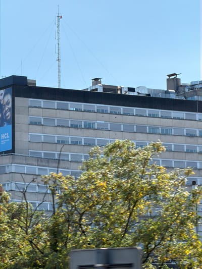 The image shows the HCL (Hospices Civils de Lyon) building in Bron, France, under a clear blue sky. The building is a multi-story concrete structure with rows of windows. A large advertisement for HCL is visible on the left side of the building. The top of the building features several ventilation systems and chimneys. A tall radio mast with antennas stands on the roof, with guy wires extending to the building. In the foreground, the blurry branches and leaves of trees with yellowing foliage suggest an autumn setting. A partial view of a vehicle is visible at the bottom of the frame.