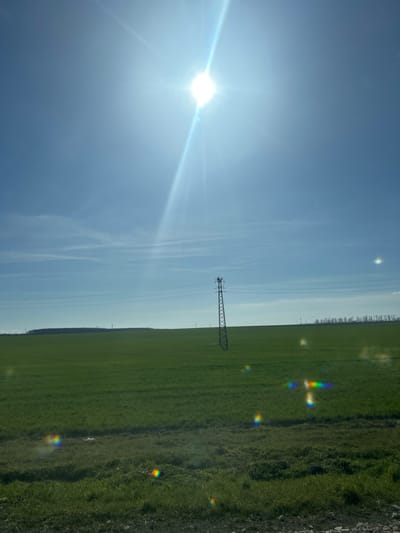 The image depicts an open, rural landscape under a clear, bright blue sky in the vicinity of Senokos, Bulgaria. A brilliant, white sun is positioned in the upper center, producing a strong diagonal sunbeam and multiple circular, rainbow-colored lens flares in the lower half of the frame. A single, dark, metallic lattice-work electricity pylon stands in the midground, slightly right of center, supporting multiple horizontal power lines that extend towards the left horizon. The ground is covered by an expansive, undulating field of vibrant green vegetation, consistent with young crops or pastureland. A distant treeline or dense foliage is visible along the far left horizon, while several faint, vertical structures resembling wind turbines are discernible on the far right horizon. The immediate foreground consists of a strip of uncultivated ground with sparse grass and exposed earth. No individuals, animals, or human actions are present within the scene.