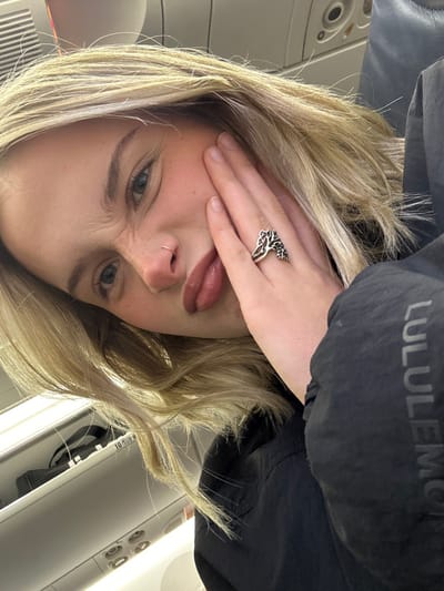 A blonde woman grimaces in an airplane seat, covering one eye with her hand. She has fair skin with freckles, blue eyes, and a nose ring. Her lips are slightly parted. She wears a large silver ring on her hand. In the background is the interior of an airplane, including a black jacket with a "LULULEMON" logo visible on the collar. The image is rotated, giving a tilted perspective.