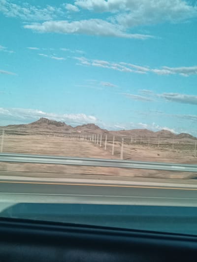 The image depicts a wide, scenic view of a desert landscape under a bright blue sky dotted with scattered white clouds. In the foreground, a highway barrier with metallic railings separates the viewer from the scene beyond. The highway itself is visible, with distinct lane markings including a yellow line. The middle ground features a flat, arid terrain, sparsely covered with dry, light-colored vegetation. A fence line, consisting of numerous vertical posts, stretches across this area, creating a visual division. In the distance, a range of rugged, rocky mountains forms the horizon, their peaks appearing somewhat weathered and eroded. The overall impression is one of a vast, open natural environment, likely in a dry climate. The time of day appears to be daytime, with bright lighting and clear skies suggesting fair weather. No people or animals are visible in the frame. The image conveys a sense of open space and natural beauty, typical of a roadside view in a remote or undeveloped region. The context provided suggests this scene is located in Kurminskoe, Kazakhstan.