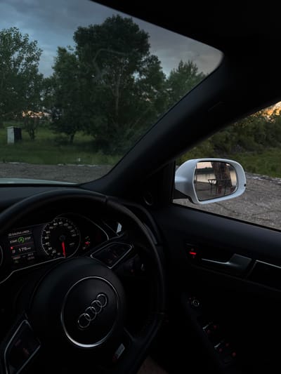 The image depicts the interior of an Audi car. The dashboard and steering wheel are prominent, with an Audi emblem visible on the steering wheel. The instrument panel indicates a speed of around 10 mph, with a mileage of 175 miles and an outside temperature of 21.5°C. Through the front windshield, trees are visible. The driver's side mirror is in view, reflecting an external scene. The location in the city of Dolni Tsibar, Bulgaria cannot be confirmed solely based on the image.