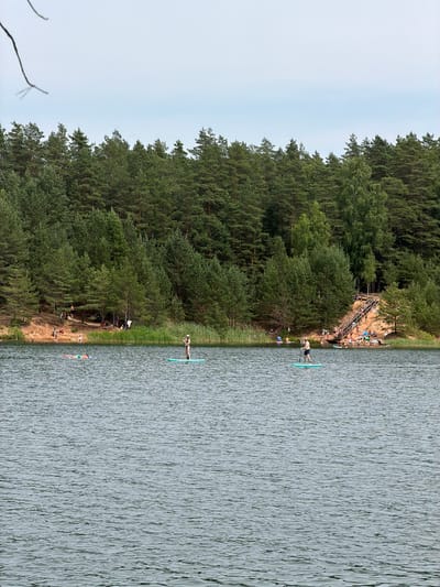 The image depicts a tranquil scene at a lake in Ikšķile, Latvia, on a bright, overcast day. The foreground is dominated by the rippling surface of the lake, with its dark, choppy waters reflecting the muted light. In the mid-ground, several individuals are engaged in water activities. Two people are standing on paddleboards, one closer to the viewer on the right, and another further to the left. A person is also seen lying on their back on the water.

The background features a sandy shore, which transitions into a dense forest of evergreen trees. Along the edge of the forest, a wooden staircase ascends a sandy incline, and numerous people are gathered on the shore and on the stairs, suggesting a popular recreational area. The overall atmosphere is one of a calm summer day at a lakeside recreational spot.