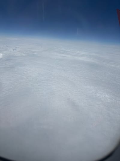 The image, taken from an airplane window, shows a vast expanse of clouds from a high altitude. The sky above is a deep, clear blue, transitioning to a lighter shade closer to the cloud layer. The clouds themselves are a uniform white, forming a textured, undulating surface that stretches to the horizon. A portion of an airplane wing, painted red and white, is visible in the upper right corner of the frame, indicating the vantage point. The scene is devoid of any urban elements, natural landscapes, or human activity, presenting a purely atmospheric view. There is no identifiable information in the image that would allow for pinpointing a specific location such as Pervomayskiy, Russia.