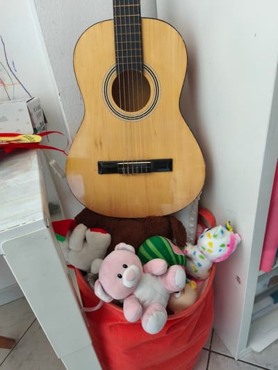 A light wood acoustic guitar is propped upright against a white wall. The guitar's body is light-colored with a dark soundhole ring and a black bridge. Its neck, also light wood, is visible with frets and tuning pegs. Below the guitar, a red fabric bin is overflowing with stuffed animals. A large brown fuzzy stuffed animal is behind a white teddy bear wearing a red and green Santa hat. A pink teddy bear is in the front, with a green and white striped stuffed object and a light-skinned doll head peeking out from behind it. The stuffed animals are crammed into the bin, some with their limbs sticking out. To the left of the guitar, a white box with a drawing of clouds and text is partially visible, along with a red ribbon with a yellow tag. The scene appears to be indoors, likely in a child's room or play area, given the stuffed toys and decorations. The lighting suggests daytime.