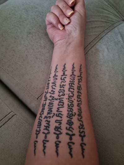 The image displays a person's left forearm and hand resting on a textured, light greenish-grey upholstered surface, likely a sofa or armchair with visible stitching. The forearm is adorned with a large, dark black tattoo composed of multiple lines of script. This script appears to be Southeast Asian, possibly a form of ancient Thai script (Sak Yant), characterized by its flowing, intricate characters. The provided OCR attempts to transcribe parts of the tattoo as "manyur WINS SY548- AJWASME SCIEN (ISMS ហ៍ប ហន្ទអរបាច់ពស لسه MSASSMS-m S&SMOSMSASS".

The person's hand is partially visible in the foreground, with fingernails painted in a pale yellow or light orange color. One nail, on the ring finger, features a small, delicate floral design in a similar light hue. The lighting suggests an indoor setting, with no direct sunlight visible. No specific activity is depicted, only the static placement of the arm. The provided context indicates the location is Xàtiva, Spain.