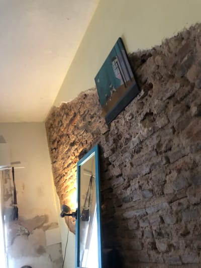 The image captures an indoor space, likely a room in Beccar, Argentina, featuring a distinctive ceiling design. The upper left portion of the ceiling is a smooth, light yellow/off-white plaster, contrasting sharply with the central and right areas which reveal rough, exposed brickwork, forming what appears to be a vaulted or arched ceiling typical of older construction. The transition between the plaster and brick is ragged, suggesting either an unfinished renovation or an intentional architectural aesthetic.
Two framed objects are prominently displayed. On the upper left, a rectangular picture with a black frame shows artwork predominantly green and blue, containing indistinct shapes and possibly unreadable text. On the upper right, a larger rectangular frame with a noticeable light blue border appears to be a mirror. This mirror reflects a brighter area, possibly a window or an opening to another room, along with parts of what looks like a white curtain or window frame, and some dark, linear objects below it. A small, dark-colored spotlight-style light fixture is attached to the exposed brick ceiling near the blue-framed mirror, casting illumination onto the textured brickwork.
The walls of the room are painted a uniform light yellow or off-white. On the lower right, the wall shows some stains, discoloration, or variations in texture. The reflection in the right-hand mirror also hints at the presence of a long, dark object, possibly a decorative rifle or similar item, positioned horizontally on the wall below the mirror. No people are visible, and no specific activity is taking place in the static scene. The presence of artificial light suggests the time of day is not evident. The overall impression is one of a room blending rustic historical elements with contemporary fixtures.