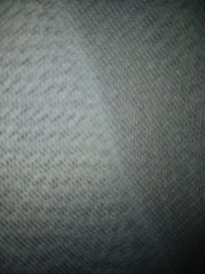 The image shows a close-up of a textile, likely denim, with a visible twill weave pattern. The fabric has a muted, cool tone, appearing as a blend of blues and grays. The texture is soft and slightly blurred, suggesting the image was taken at a close range and potentially out of focus. The lighting is uneven, with darker areas fading to lighter areas.