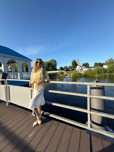 The image depicts a bright, sunny outdoor scene set on a wooden pier, described as being in Dumino, Russia. A woman with long, wavy blonde hair and round sunglasses stands in the foreground, leaning with her left arm on a grey metal railing that borders a body of dark blue water. She is smiling towards the camera. She wears a yellow and white gingham button-down shirt tied at the waist over a white tank top, paired with a white tiered midi skirt and light-colored flat sandals with ankle straps.
The pier is constructed from wide, brown composite decking planks. To the left, a white gazebo-like structure with a blue, sloped roof is partially visible. A black bucket or container rests on the pier near the gazebo.
Across the body of water, a prominent white architectural complex, characteristic of a monastery with multiple domes (some green, some dark/silver) and bell towers, is situated on the far bank. This complex is surrounded by lush green trees and bushes. Several small, indistinct figures, including what appear to be children, are visible on the distant bank near the water's edge. Approximately ten dark-colored ducks are scattered and swimming in the water between the pier and the far bank. The sky above is clear and bright blue, indicating fair weather. Strong sunlight casts distinct shadows from the railing onto the pier.