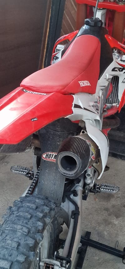 A red and white Honda CRF dirt motorcycle is observed stationary, positioned on a black metal bike stand. The motorcycle is presented from a three-quarter rear perspective. Its seat is red and textured, bearing the "HONDA" logo. A black fuel tank cap is visible at the top of the tank area. The rear fender is red with an "O'NEAL" logo. White side panels feature "CRF" branding, with the rear right panel displaying notable damage and cracking. A carbon fiber exhaust muffler with a circular opening is prominently located on the right side. Below the muffler, a black component is adorned with a "HOT RODS" sticker. Serrated metal footpegs are visible on both sides of the frame. The drive chain is present on the left side of the rear wheel, which is fitted with a knobby "PIRELLI" dirt tire. The ground directly beneath the motorcycle is a dusty concrete or paved surface. The background consists of a blurred wall, potentially brick or wood, with a dark vertical element that may be a door frame or support beam. The lighting suggests ambient daylight. The scene is located in Kolarovo, Bulgaria.