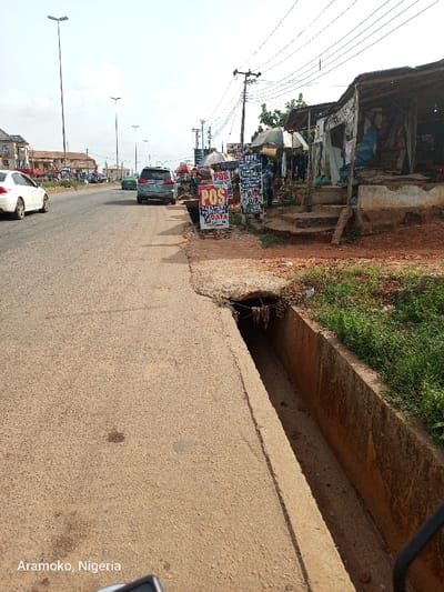 The image shows a street scene in Ado Ekiti, Nigeria, at coordinates 7.64 latitude and 5.221 longitude. The scene features a paved road with a gutter running along the side. Several cars are visible on the road, including a silver sedan parked on the right side. There are various signs on the side of the road including one that says "POS Passport Photocopy Data." There are also several small businesses along the street, including one that appears to be selling clothes. The street has a few power lines running above it. There is a large, open drainage ditch in the foreground. There are a few people in the background. The sky is clear and the weather appears to be sunny. There are trees and plants on either side of the road. The road is made of asphalt and the gutter is made of concrete.