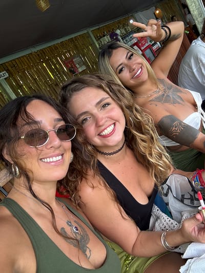 Three smiling women are captured in a selfie. The woman at the top left wears a white top and gives a peace sign. The woman in the middle has curly hair and a black top. The woman at the bottom wears sunglasses and a green top, revealing a tattoo. The group is indoors. A wooden wall is in the background.