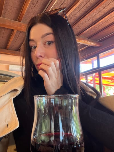 A medium-shot image depicts a female subject seated indoors in Civitavecchia, Italy. The subject, with long dark hair, dark eyebrows, and brown eyes, is looking directly forward. Dark-framed sunglasses are perched on top of her head. Her left hand is raised to her chin/mouth area, with fingers slightly curled. She is wearing a black top or sweater, and a light beige or tan collared jacket is draped over her shoulders. In the lower foreground, a clear glass containing a quantity of red liquid, consistent with red wine, is positioned. The interior background features an exposed wooden beam ceiling with light-colored panels. To the right, windows reveal an outdoor area with strong sunlight, red/orange colored structures, and green foliage. An air conditioning unit is partially visible on a light-colored wall in the upper left background.