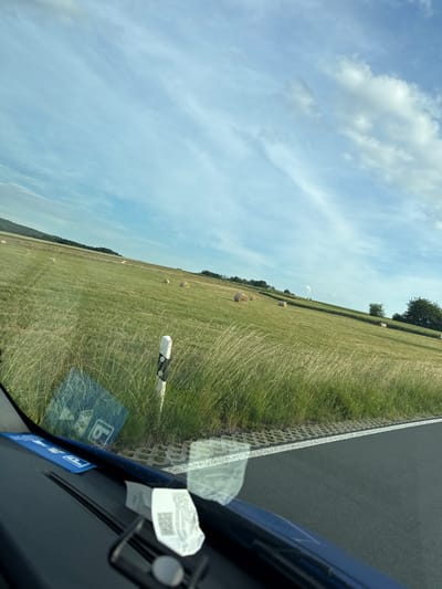 The image, taken from inside a vehicle, displays a rural landscape. In the foreground, the dark dashboard of the car is visible, with a white slip of paper featuring a QR code resting on it. A blue parking permit is also affixed to the windshield. The view through the windshield reveals a vast, green field dotted with numerous round hay bales, indicating a recent harvest. A road runs along the right side of the frame, bordered by a grey concrete barrier and a white line. A white and black striped post stands near the edge of the field. In the background, rolling hills covered in trees form a distant horizon under a partly cloudy sky. The overall scene suggests a drive through the countryside near Niederwetz, Germany.