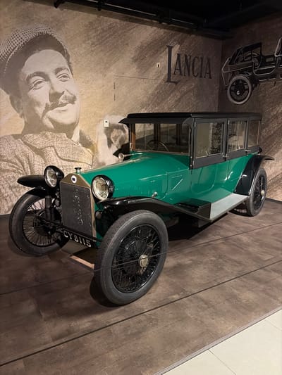 This media file depicts a vintage dark green Lancia automobile displayed in an indoor setting, likely a museum or exhibition in Wassenaar, Netherlands. The classic car, an early 20th-century model, features a dark green body, black fenders, and a black-framed cabin with windows. Its front is equipped with two prominent round headlights, a metallic mesh radiator grille, and a license plate clearly displaying "CY8115". The car rides on spoked wheels fitted with black tires, and a spare tire is mounted on its side.
The background consists of a dark, textured floor and a large wall adorned with graphic prints. Dominating a section of the wall is the word "LANCIA" rendered in large, dark serif letters, identifying the vehicle's manufacturer. Adjacent to the car's front is a striking, sepia-toned portrait of a man, possibly Vincenzo Lancia, the company's founder. He is depicted with a thoughtful expression, looking upwards, and wearing a distinct cap and textured clothing. Above the Lancia text, a line-art illustration of an early automobile's chassis or open-wheel design is also visible. The scene suggests a static exhibit, showcasing automotive history.