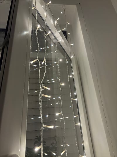 The image shows a close-up view of a string of white LED lights hanging vertically along a white window frame. The lights are spaced at intervals and are illuminated, casting a warm glow. The window appears to be a double-glazed unit with blinds or shutters visible through the glass. The scene is indoors, likely a residential or commercial interior, and the time of day is indeterminate due to the artificial lighting and reflections. There is no identifiable location in Münster, Germany, that can be determined from the visual information provided. The image focuses solely on the decorative lighting and the window structure.