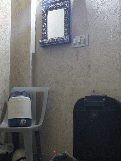 The image shows a cluttered indoor scene, possibly a residential space in Abbottabad, Pakistan. The dominant elements are a textured wall with a subtle patterned wallpaper, a black object that appears to be a water heater or some kind of appliance mounted on the wall, and a decorative mirror with an ornate frame hanging on the wall. Below the mirror, there is a light switch panel with multiple switches.
In the foreground, a white plastic chair is partially visible. On the chair, there is a white cylindrical object with a blue band, likely a water filter or purifier, and a small nozzle or spigot. The background of the image is dark and indistinct, but a soccer ball and other unidentifiable objects can be seen.
The lighting suggests it could be daytime, but the overall ambiance is dim, with shadows casting a somewhat mysterious or neglected feel. There is no visible text in the image. The scene appears to be a candid snapshot of a domestic setting, focusing on everyday objects and the textures of the environment.