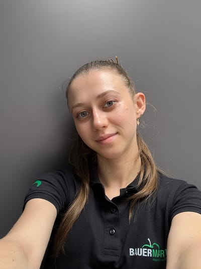 This media file is a selfie of a young woman with long, light brown hair. She is wearing a black polo shirt with a green logo that reads "BAUER MART" and "Regionaler Dienst". A smaller green text below it is partially obscured. The woman is looking directly at the camera with a slight smile. Her left eye is a light blue, and her skin appears clear. A small gold earring is visible in her left ear. The background is a plain, dark gray surface. The image is oriented sideways. The lighting suggests an indoor setting, possibly during the day. There are no explicit cues about the weather or specific location within Elsenfeld, Germany, beyond the text on her shirt.