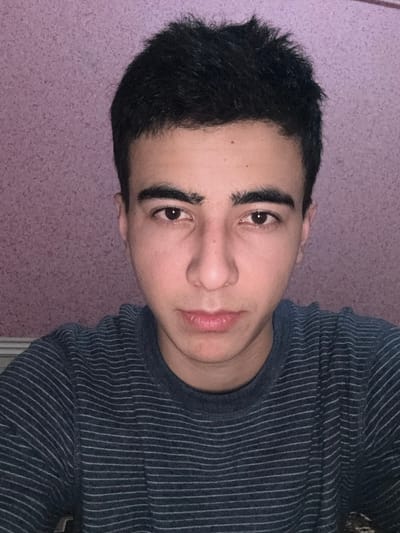 A close-up of a young man with short, dark hair and defined eyebrows. He wears a striped long-sleeved shirt. His skin is smooth and fair. The background is a textured purple wall. The image is rotated, causing the subject to appear sideways. His expression is neutral, and the lighting is soft and slightly muted.