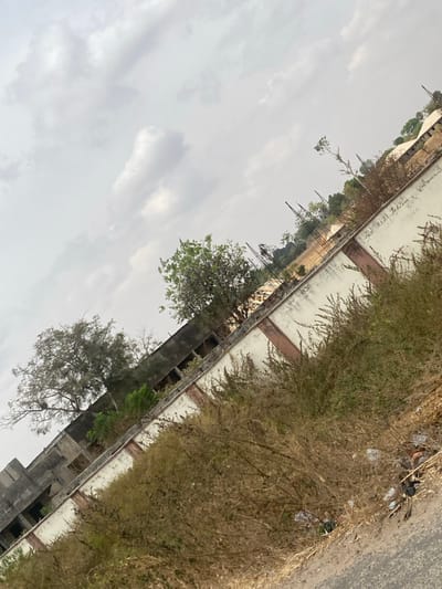 The image captures an outdoor scene in Angwan Eggon, Nigeria, taken from an oblique angle tilted upwards to the right. The foreground presents a section of dark asphalt or dirt road, bordered by an unkempt area of dry, light brown, and green vegetation. Scattered white plastic bags and other debris are visible within this vegetation near the road. A light-colored, possibly cream-painted, concrete or masonry wall with vertical reddish-brown accents runs diagonally across the midground. Behind the left portion of this wall, a dilapidated, multi-story building with dark, visible window openings is present. To the right, behind the wall, several vertical metal rods, consistent with rebar for construction, protrude upwards. Various trees with green foliage are distributed across the mid-to-background, and distant structures with light-colored roofs are faintly discernible. The sky is uniformly overcast with shades of grey and white clouds. No individuals are depicted in the scene.