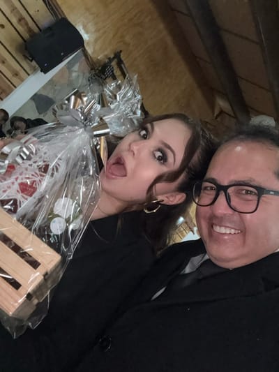 A woman and a man in black suits pose for a selfie. The woman, with brown hair and makeup, holds a gift basket wrapped in clear plastic. The man wears glasses and smiles. The background features wood paneling, a speaker, and what appears to be attic or storage space. The scene suggests an event, potentially a celebration or gathering.