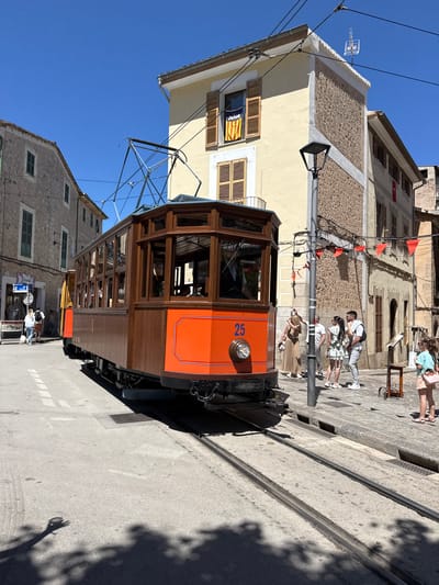 The image shows a brown and orange tram labeled with the number 25 on its front, traveling on tracks in Sóller, Spain. Several people are walking along the street adjacent to the tram tracks. Buildings with stone and yellow exteriors line the street. Orange flags are strung between buildings. Overhead power lines run above the tram. The sky is clear and blue.