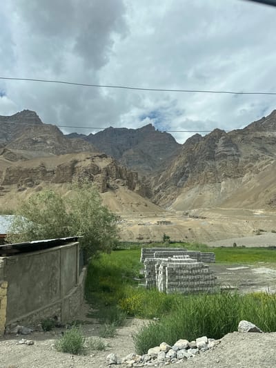 A wide shot captures a stark, mountainous landscape under a cloudy sky in Mulbekh, India. In the foreground, a rough, gray stone wall runs along the left side of the image, partially obscured by a weathered concrete structure with a corrugated metal roof. To the right of the wall, a lush green field is dotted with small yellow wildflowers. In the mid-ground, stacks of light gray bricks are neatly piled on the grass, suggesting construction or storage. Beyond the field, the terrain rises dramatically into barren, layered mountains with tan and brown hues. A few scattered buildings and a dirt road are visible in the valley between the foreground and the mountains. Two thin black wires stretch horizontally across the upper portion of the image, suggesting the presence of power lines. The overall scene is one of rugged natural beauty with hints of human activity in a remote, high-altitude environment.