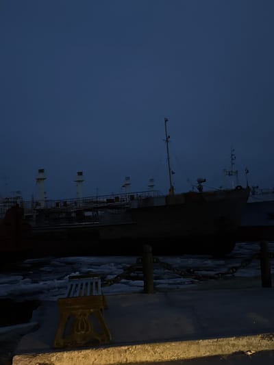 A long ship is docked, viewed from ground level at twilight. The ship is dark, with a complex superstructure and a tall mast. A concrete barrier is on the right, with a decorative gold mooring cleat. The sky is a uniform dark blue. Based on the presence of ships and docking facilities, the location might be in or near the city of Zhatay, Russia, given its location on the Lena River which hosts shipping activity. No people are visible.
