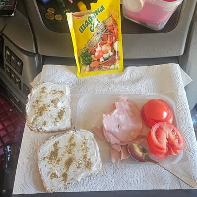 The image depicts a simple meal laid out on a white paper towel, likely inside a vehicle, as suggested by the dashboard controls visible in the background. Two slices of bread, spread with a white creamy substance and sprinkled with a green herb mixture, are positioned on the left side of the frame. To the right, a clear plastic container holds several slices of mortadella and two sliced tomatoes. A silver spoon rests next to the mortadella and tomatoes. In the background, a yellow packet of spice mix is visible, along with a few coins. The overall scene suggests a meal taken on the go, possibly by a traveler. The location is identified as Mas Aliu, Spain.