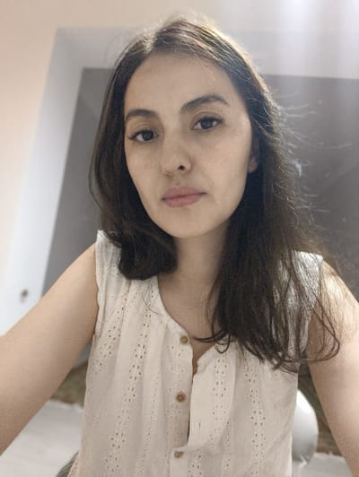 This is a medium close-up selfie of a young woman indoors, likely in a domestic setting. She has dark brown hair, parted to the side and falling past her shoulders. Her eyebrows are dark and well-defined, and she is wearing subtle eye makeup. Her lips are a natural pinkish hue, and her skin has a few light freckles visible. She is wearing a white, sleeveless top with a delicate floral eyelet pattern and small wooden buttons down the front. The background is softly blurred, but hints of a room with light-colored walls and possibly a mirror or large reflective surface are discernible. The lighting appears to be natural, suggesting daytime. The woman's expression is neutral, and she is looking directly at the camera. There is no visible text in the image. The overall impression is that of a casual, unposed moment captured by the subject herself.