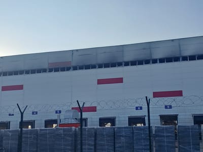 The image shows the exterior of a large, white industrial building with a grey roof. The building has several large, red rectangular markings on its facade. Along the base of the building, there are multiple loading bay doors, some of which are open, revealing dimly lit interiors. A wire fence with barbed wire on top runs along the front of the building, separating it from the viewer. Several numbered signs, ranging from 4 to 9, are visible on the fence posts. The sky above is clear and light blue, suggesting daylight. The overall scene depicts a commercial or industrial facility in Plastik, Russia, with signs of activity around the loading bays.
