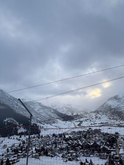 A snowy mountain scene under an overcast sky. Several power lines traverse the landscape. In the distance, a break in the clouds allows sunlight to peek through. The right side of the image contains a small town covered in snow, with buildings clustered together. A safety net, likely belonging to a viewing platform, is visible in the lower right.