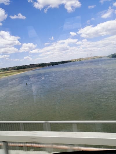 The image shows a wide river with calm water under a blue sky with scattered white clouds. The riverbanks are lined with green vegetation, and a few power pylons are visible in the distance on the left side of the river. In the foreground, a white railing of a bridge or elevated walkway is visible, suggesting the photo was taken from an elevated position. There is a single green buoy floating in the river. A barge is visible on the far right bank of the river. The scene is likely in or near Stelnica, Romania, given the mention of the location.