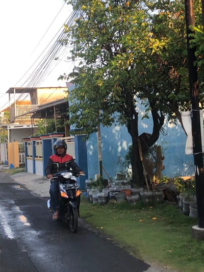 A person wearing a black and red jacket and a black helmet is riding a black motorcycle with a license plate reading "W 4798 RJ" and "06-23" in Kalidawir, Indonesia. The motorcycle has its headlights on and the rear turn signals are illuminated. The rider is wearing blue jeans and flip-flops. The motorcycle is on a wet asphalt road. To the right of the road is a grassy area with potted plants and some fallen leaves. Behind the grass is a blue wall with trees casting shadows. In the background, there is a building with an elevated porch and a light-colored roof, as well as power lines. The scene appears to be a street in a residential area during daylight.