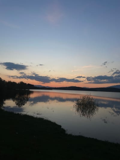 The image shows a landscape at dusk, likely in Domlyan, Bulgaria. A body of water, potentially a lake, reflects the sky and surrounding land. The sky transitions from a light blue to shades of orange and pink near the horizon, with scattered clouds. A mountain range or hills are visible in the distance. The reflection creates a mirror-like effect on the water's surface. Dark vegetation, likely grass and trees, occupies the foreground. A small, bushy tree grows near the water's edge. A thin crescent moon is visible in the upper left of the sky. There are no people present in the image.