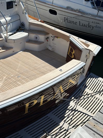 An eye-level, medium shot captures the stern of a dark-hulled yacht docked in the Village of Sag Harbor, United States of America, on a sunny day. The yacht's name, "PLAN" in large, golden, serif lettering, is emblazoned on its hull, partially obscured by reflections. Below this, the word "JUPITER" is visible in smaller, golden lettering. The yacht features a teak deck with a textured, slatted wooden platform extending from the stern. A white cushion sits on a built-in bench to the left of the frame, adjacent to stainless steel railings. A coiled white hose and a faucet are visible on a step leading to the deck. The dark water of the harbor is visible to the right. Another white yacht, with the name "Plane Lucky dos" and "SUN ODYSSEY 45 DS" written on its hull, is moored alongside. The scene is brightly lit, with distinct shadows cast by the yacht's structures and railings across the deck.