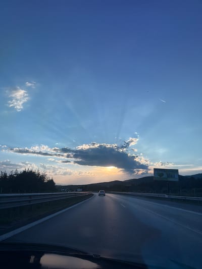 The image depicts a view from inside a vehicle driving on a highway. The sky is a clear blue with dramatic sunbeams radiating from behind a large cloud formation. The sun is setting, casting a warm glow on the horizon. In the distance, mountains are visible, silhouetted against the sky. There are trees lining the side of the road, and a billboard with Cyrillic text, likely advertising "TERMO POMPI" (heat pumps), is also present. A single car is visible on the highway ahead, and distant trucks can be seen in the far background. The highway appears to be in a rural or semi-rural setting, possibly in or near Eleshnitsa, Bulgaria, given the presence of Cyrillic text on the billboard and the mountainous terrain. The reflection of the dashboard and windshield is visible in the foreground.