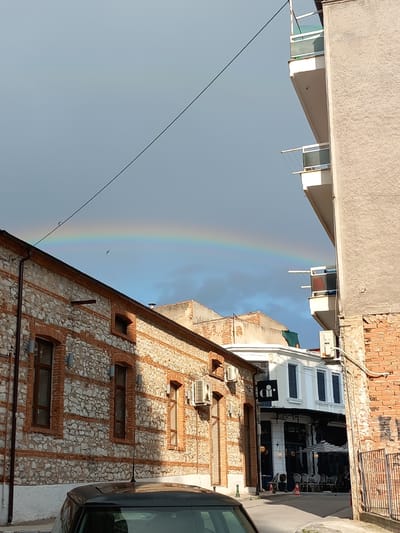 The image shows a partial rainbow in the sky over an urban scene in Drama, Greece. The view is angled upwards, with buildings framing the rainbow. One building has a facade of light-colored stone with brick accents around the windows and in vertical strips. There are air conditioning units mounted on its exterior. To the right of this building is a glimpse of a modern, white building with a black awning over an outdoor dining area; tables and chairs are visible. Part of a dark-colored car is visible on the right edge of the frame. Electrical wires cut across the sky near the rainbow. The sky is partly cloudy. The architecture suggests a mixture of older and more contemporary styles.