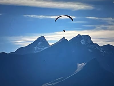 The image depicts a paraglider in flight above a mountain range. The paraglider is silhouetted against a bright blue sky with scattered white clouds. Below the paraglider, several snow-capped mountain peaks are visible, their outlines dark against the sky. The foreground is a dark blue expanse, likely representing a valley or lower elevation. The image is taken from a distance, capturing the vastness of the landscape and the smallness of the paraglider within it. The scene is tranquil and offers a sense of freedom and adventure.