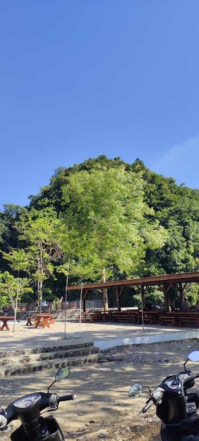 The image depicts an outdoor seating area with wooden picnic tables and benches under a covered pavilion, situated at the base of a lush, tree-covered hill. The sky is a clear blue. In the foreground, a black motorcycle is parked. The location is identified as Lamseunia, Indonesia. The scene appears tranquil, with no apparent activity or interaction among people, if any are present. The environment is natural and scenic, with a focus on the vegetation and outdoor amenities.