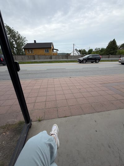 The image captures a view from a bus stop in Sarpsborg, Norway, looking out towards a road with traffic. A person is seated at the bus stop, with their legs and feet visible in the foreground, wearing light blue pants and white sneakers. The bus stop structure's black metal frame is visible on the left side of the image. Beyond the bus stop is a sidewalk paved with red square tiles, followed by a paved road where a black Volvo SUV and another car are driving. A wooden fence runs along the roadside, separating it from a residential area with a yellow house and trees. The sky is overcast and cloudy.