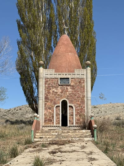 This image depicts a small, distinctive memorial structure situated in an outdoor, semi-arid setting in Barbulak, Kyrgyzstan. The building features a reddish-brown conical roof, possibly made of terracotta, and walls constructed from irregular reddish-brown stones set with white mortar. White brickwork accents the facade, particularly beneath the cornice and surrounding a dark inscription plaque.
Two decorative pillars, topped with sphere-like elements, frame a small, arched entrance. Greenish accents are visible at the base of these pillars and along the concrete steps leading to the entrance. To the left of the structure, a large, leafy tree stands prominently against a clear blue sky, which shows faint contrails. The background to the right consists of a dry, light-colored hillside covered with sparse, yellowish-green grass and some bare vegetation.
A dark plaque above the entrance bears Cyrillic script, which reads:
"АЛАЧАЕВ" (ALACHAEV)
"БААТЫРКАН" (BAATYRKAN)
"1840 / 1929"
This text indicates the structure is a memorial to Baatyrkan Alachaev, who lived from 1840 to 1929. The image is taken in bright daylight under a clear, sunny sky, with no human activity visible. The overall scene suggests a place of commemoration or historical significance within its natural, dry landscape.